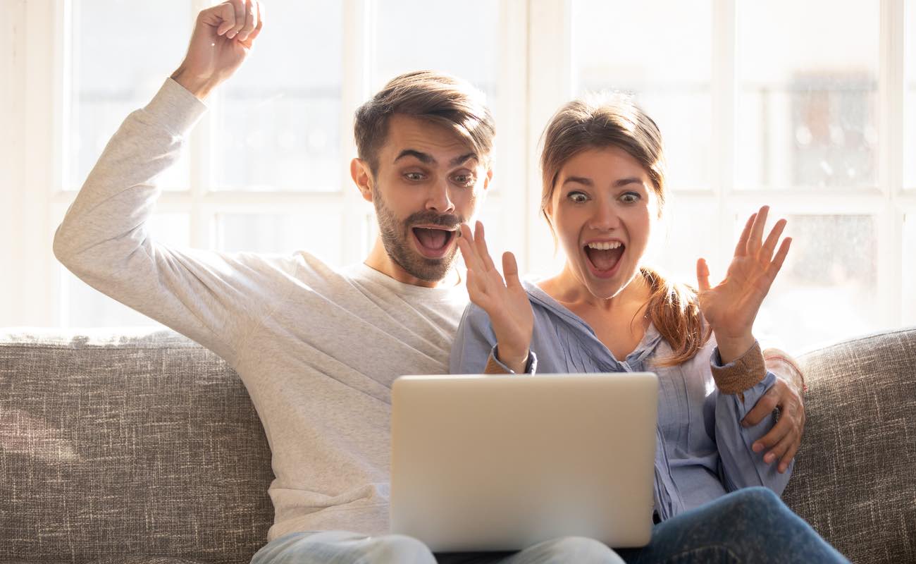Couple sitting on couch using laptop looking at device screen feeling triumph from online win