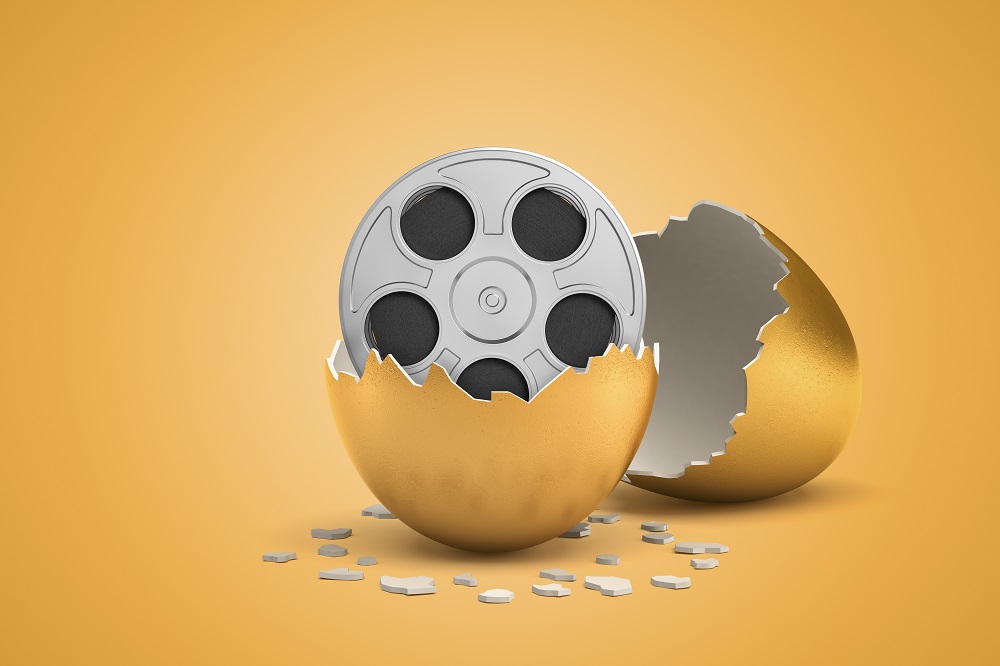 3d rendering of broken in two golden egg with retro film reel inside on light ocher background. - stock photo
