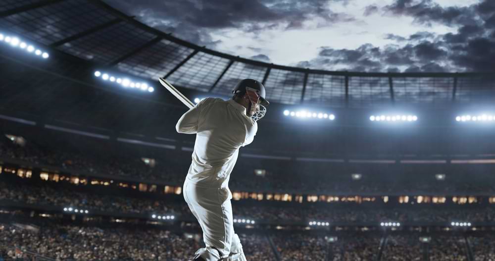 Cricket player in full protective gear swinging a bat under bright stadium lights during a night match with a packed crowd in the background.