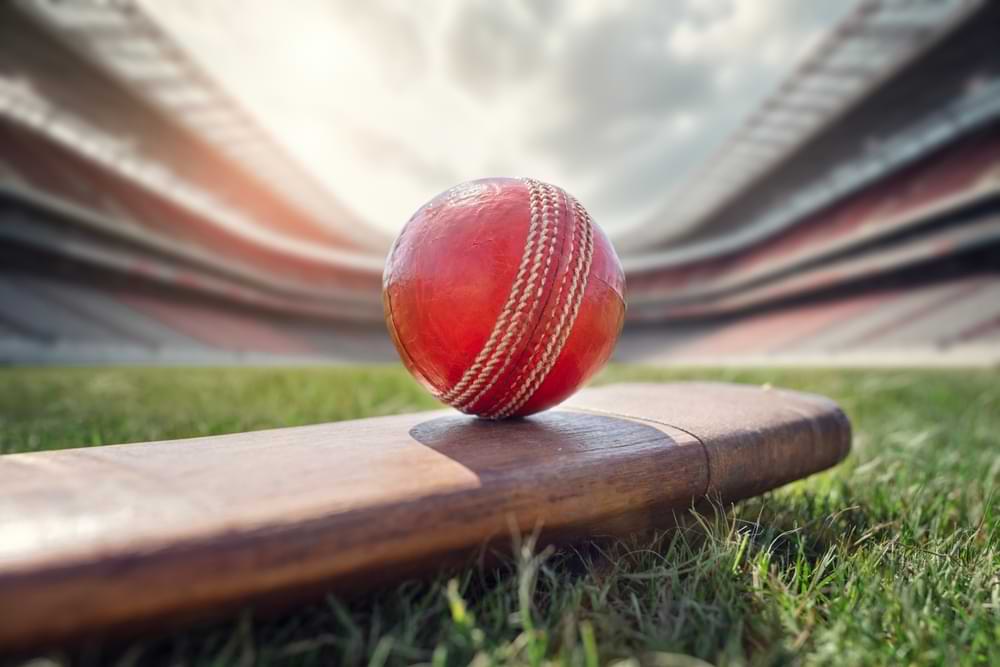 Red cricket ball resting on a cricket bat on a grassy field inside a large stadium.