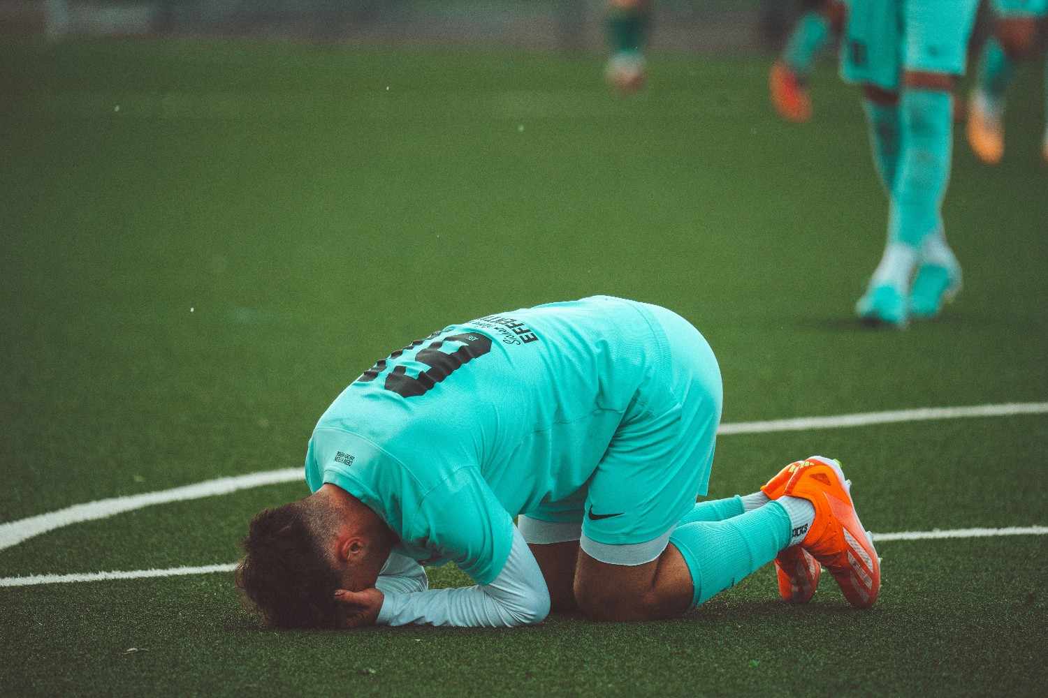 Footballer in a turquoise kit collapses to both knees on the pitch, head pressed to the turf near a white field line, bright orange boots striking against the green grass.